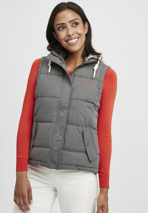 Un gilet matelassé gris avec une capuche amovible, des poches avant et des accents à cordon de serrage ; porté sur une chemise à manches longues rouge et un pantalon crème.