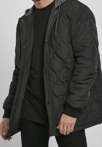 Homme portant une veste matelassée noire avec des boutons-pression et des poignets côtelés sur une chemise noire, debout devant un fond uni.