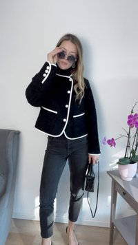 Casaco de bouclé preto com detalhes brancos, formato rectangular e botões redondos, combinado com jeans escuros e uma bolsa preta. Cadeira cinza e orquídeas cor-de-rosa ao fundo.