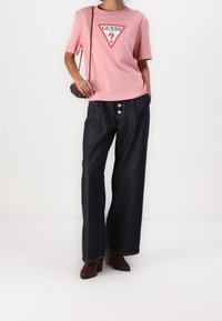 Mujer con una camiseta rosa de Guess, jeans de pierna ancha oscuros con detalles de botones, botas de tobillo marrones y un pequeño bolso cruzado marrón.