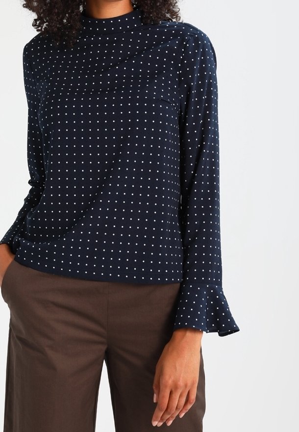 Donna che indossa un top blu navy a maniche lunghe con pois bianchi, con polsini arricciati e pantaloni marroni, in piedi con una mano in tasca.