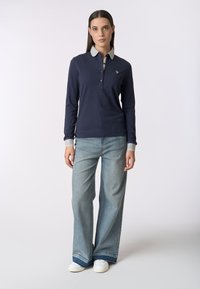 Maglietta polo a maniche lunghe blu navy con colletto grigio chiaro, due bottoni e logo ricamato. Abbinata a jeans chiari a gamba larga e scarpe bianche.