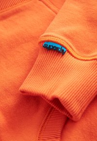 Narančasti rebrasti manžet majice s plavom tkaninom oznakama s natpisom «superdry» ušiven na šav.