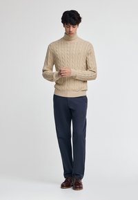 Beige kabelstickad polotröja med ribbad fåll, tillsammans med marinblå byxor och bruna snörskor. Den enkla designen framhäver texturen.