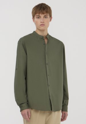 Camicia verde oliva a maniche lunghe con colletto alla mandarino, chiusura frontale con bottoni, texture liscia e leggero lucido; abbinata a pantaloni beige.