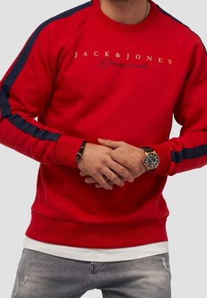 Sweat-shirt rouge avec des rayures latérales bleu marine, présentant le texte "JACK & JONES Originals". Tissu doux, bords côtes aux poignets et à l'ourlet. Porté avec un jean gris.