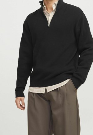 Hombre con suéter negro con cremallera hasta el pecho sobre camisa beige a cuadros, pantalones marrones plisados, con la mano en el bolsillo, sobre fondo liso.
