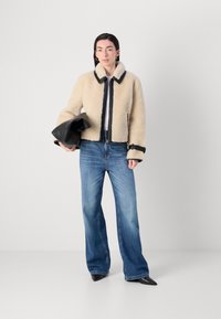 Crème kleurige faux schapenwollen jas met zwarte afwerking, gecombineerd met lichtblauwe wijde jeans en zwarte puntige schoenen. Leren tas onder de arm.