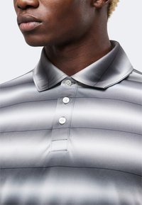 Κοντινό πλάνο ενός ατόμου που φορά μια γκρι ριγέ πόλο μπλούζα με διπλωμένο γιακά και τρία λευκά κουμπιά κλεισμένα στο λαιμό.