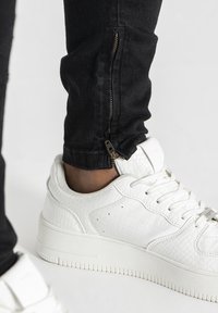 Biele textúrované tenisky s krokodílou vzorom, skombinované s čiernymi slim-fit džínsami s detailom bočného zipsu na členku.