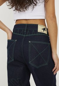 Dark blue denim pants with green stitching, featuring unique pocket designs and a cream label with a star logo on the back waistband.