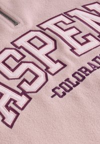Fleece pulóver, világos rózsaszín színben, nagy, lila hímzett "ASPEN" felirattal rózsaszín kiegészítőkkel, félig cipzáros gallérral a záródásért.
