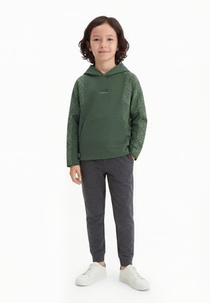 Dziecko z włosami do ramion, ubrane w zieloną wzorzystą bluzę z kapturem, ciemnoszare spodnie dresowe i białe sneakersy, stoi z lekkim uśmiechem.