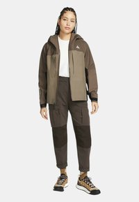 Brun vattentät jacka med två framfickor, matchad med mörka cargobyxor. Outfiten har sportiga skor med varierande texturer och färger.