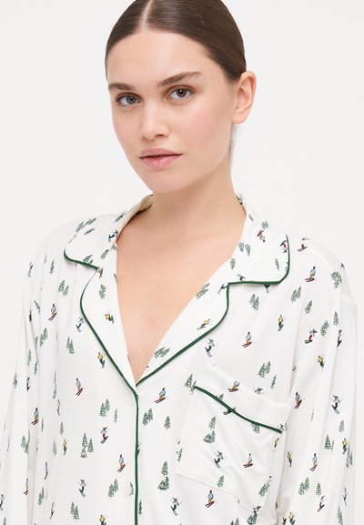 Λευκό πυτζάμα με μακριά μανίκια, με πράσινη διακόσμηση και εκτύπωση με θεματολογία σκι που απεικονίζει σκιέρ και δέντρα. Διαθέτει τσέπη στα αριστερά του στήθους.