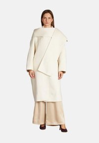 Cappotto di lana bianco con un design asimmetrico, maniche larghe e un grande colletto. Abbinato a pantaloni in satin beige e tacchi scuri.