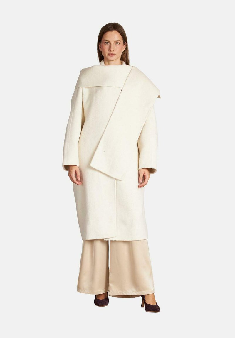 Cappotto di lana bianco con un design asimmetrico, maniche larghe e un grande colletto. Abbinato a pantaloni in satin beige e tacchi scuri.