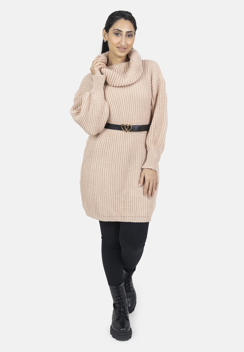 Suéter de punto oversized en rosa claro con cuello de cisne, ceñido por un cinturón negro con hebilla en forma de corazón, combinado con leggings negros y botas.