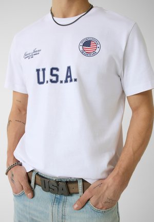 Persoon draagt een wit T-shirt met de tekst "U.S.A.", Amerikaans vlagembleem, "Guess Jeans" logo, lichtblauwe spijkerbroek en een bruine riem met "USA" gesp.