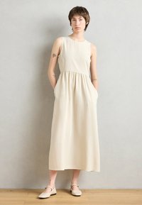 Sleeveless, cream-colored maxi dress with a gathered waist, a relaxed fit, and side pockets, paired with light-colored flats.