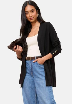 Žena s dlouhými tmavými vlasy je oblečena v černém cardigan s zlatými knoflíky, bílém tílku, modrých džínách a hnědém pásku, drží složenou hnědou tašku.