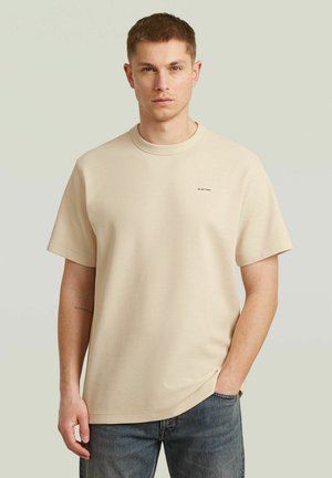 Junger Mann mit kurzem Haar, der ein beiges, strukturiertes T-Shirt mit kurzen Ärmeln und blaue Jeans trägt, steht vor einem schlichten hellen Hintergrund.