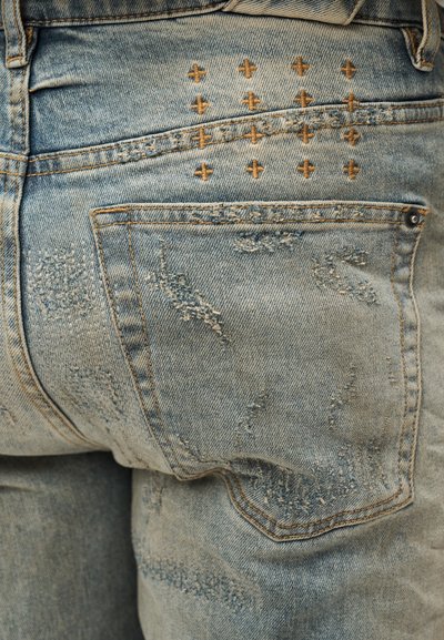 Hele sinise teksapüksid, millel on kulunud laigud, õrn tuhmumine ja kuldse õmbluse aktsendid tagatasku kohal.