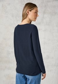Marineblaues langärmeliges, strukturiertes Oberteil mit Rundhalsausschnitt und lockerer Passform, kombiniert mit hellblauen Jeans. Die Rückansicht hebt die Details an Schulter und Ärmeln hervor.