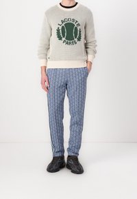 Stickad krämfärgad tröja med grön Lacoste-logotyp, ihop med blå mönstrade byxor med vita sidostripes och svarta klumpiga sneakers.