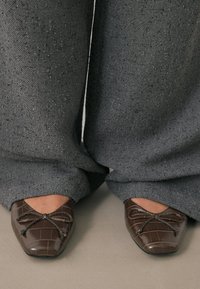 Ballerines en cuir verni marron avec un bout pointu, présentant un motif crocodile texturé et un détail de nœud décoratif. Associées à un pantalon gris à jambes larges.