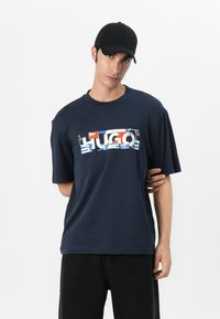 Navy blå oversized t-skjorte laget av bomull, med et flerfarget "HUGO"-grafikk over brystet. Svart justerbar caps, båret i en lett skrå vinkel.