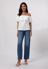 Weißes schulterfreies Oberteil mit geschichteter Rüschendesign, strukturiertem Stoff, kombiniert mit hochgeschnittenen blauen weit geschnittenen Jeans und weißen Riemchenschuhen.