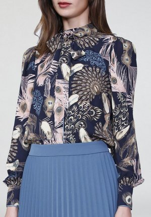 Blouse bleu marine avec imprimé plumes de paon, dotée d'un col à nouer et de manches longues, associée à une jupe plissée bleu ciel. Texture de tissu lisse.