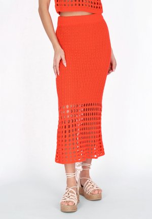 Vrouw die een feloranje gebreide midirok draagt met een doorschijnende gehaakte zoom en beige plateausandalen met enkelriempjes.