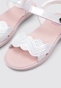 Sandalia blanca con un diseño de correas onduladas y costuras elevadas, plantilla de color rosa suave y suela de goma rosa. Correa ajustable en el tobillo.