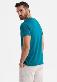 Teal cotton t-shirt with a round neckline and short sleeves, worn by a man with hands in pockets, featuring a minimal design.
