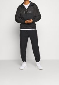 Windbreakerjack met een half-rits aan de voorkant en een logo in het zwart, gecombineerd met zwarte joggers. Beide hebben elastische manchetten en aansnoer opties.