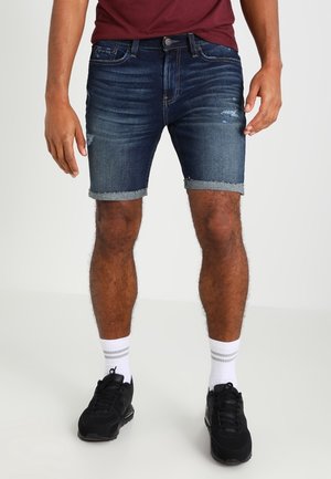 Denimshorts i mørk vask med slidte detaljer, opsmøgede kanter, lukket med knap og lynlås, stylet med en mørkerød skjorte og sorte sneakers.