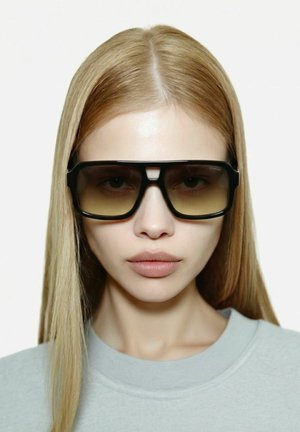 Jeune femme aux cheveux blonds raides portant des lunettes de soleil surdimensionnées rectangulaires dégradées marron et un haut gris clair à col rond.