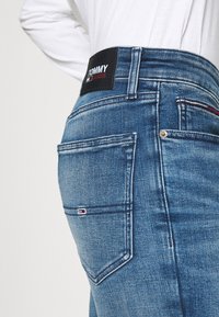 Denim jeans in medium blue with a slightly distressed finish, featuring a black Tommy Jeans logo patch and red accent stitching on the back pocket.