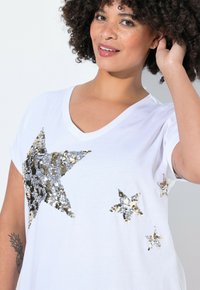 Bílé tričko s výstřihem do V a krátkým rukávem, zdobené zlatými a stříbrnými flitrovými hvězdami různých velikostí na přední straně.