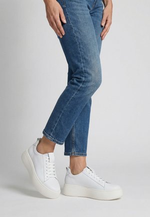 Person trägt weiße Plateau-Sneaker und blaue kurze Jeans, steht vor einem schlichten hellen Hintergrund.