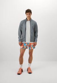 Chaqueta atlética gris, camiseta blanca, pantalones cortos multicolores con patrones naranjas y azules, y zapatillas de correr rosas con acentos negros.