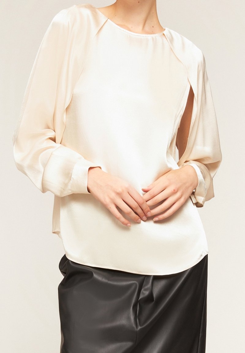 Femme portant un chemisier en satin crème aux manches fendues ouvertes et un pantalon en cuir noir, les mains délicatement jointes à la taille.