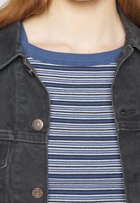 Veste en denim noir avec des boutons en cuivre, portée sur un haut en tricot bleu et blanc à rayures, doté d'un col rond et d'une texture côtelée.