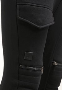 Svarta joggingbyxor med matt yta, utrustade med en ficka med dragkedja och en platt lap med "BLACK KAVAR"-logotyp på låret.