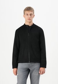 Joven con una chaqueta negra con cremallera y cuello de banda, y jeans gris claro, de pie contra un fondo blanco.