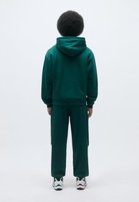 Felpa verde con cappuccio regolabile, abbinata a pantaloni jogger verdi coordinati. Presenta una texture liscia e una vestibilità comoda. Scarpe da ginnastica bianche visibili.