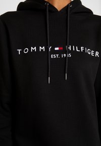 Schwarzer Hoodie aus Baumwolle mit strukturierter Oberfläche. Verfügt über weiße, gestickte Markenkennzeichnung und ein kleines, farbiges Logo; silberne Beschläge an den Kordeln.