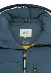 Pufferjakke med lynlås foran, mørkeblå overflade, gul foring og en logoetiket indeni. Har en struktureret stof og høj krave.